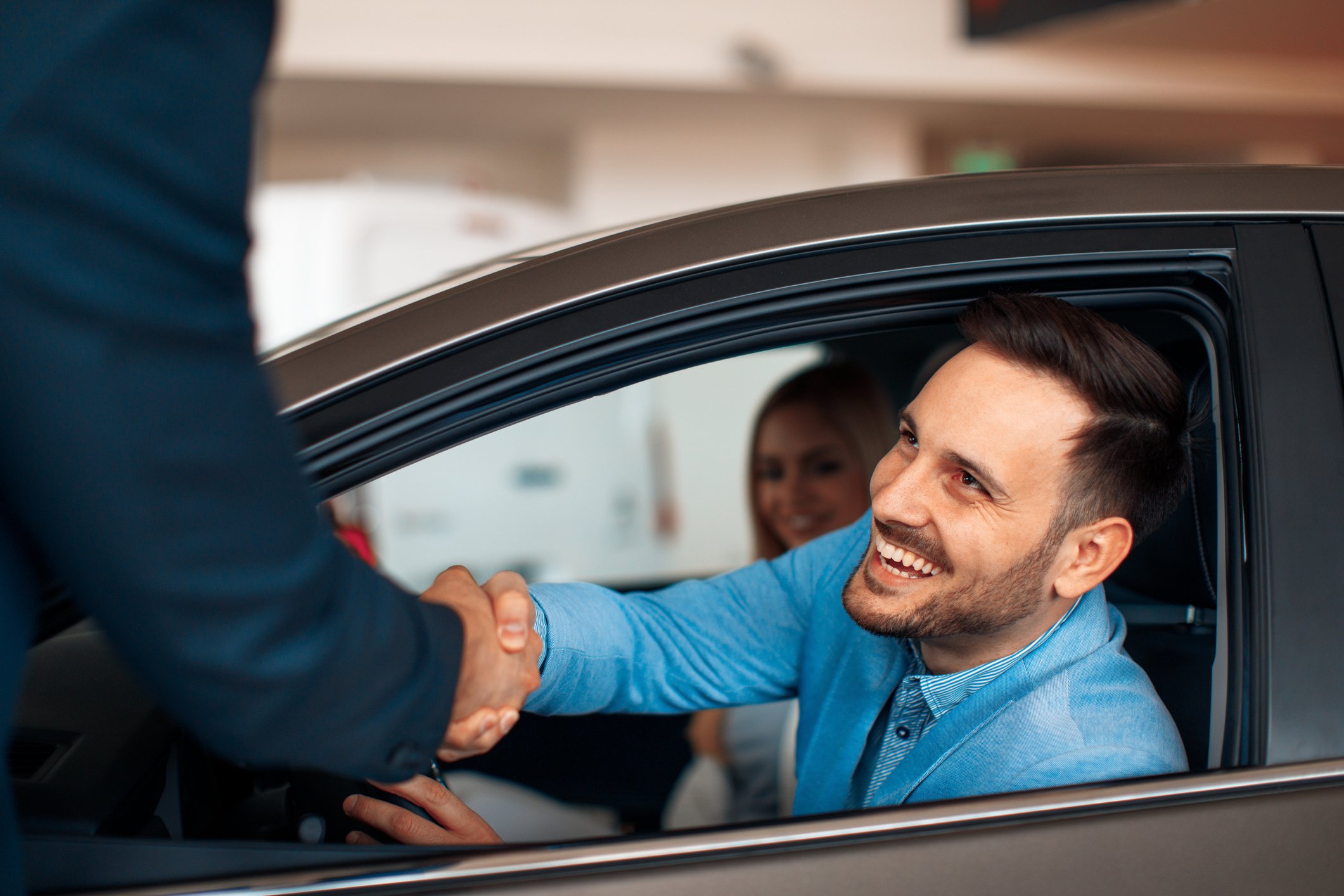 A young, smiling man shaking hands with a car salesman while still inside a car he's buying with his wife.