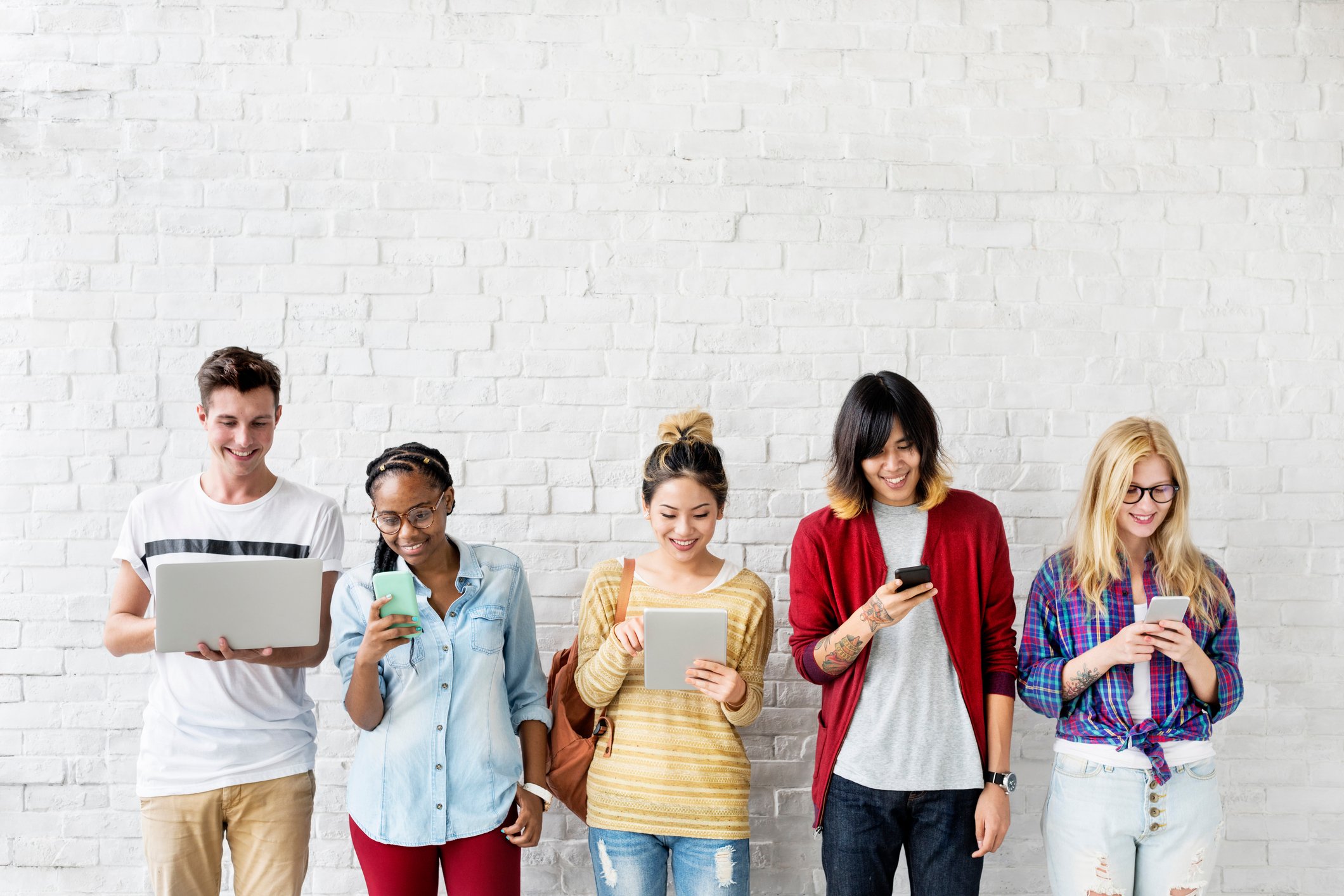 Five young people lined up against a white brick wall while looking at their smartphones, tablets, and laptops.