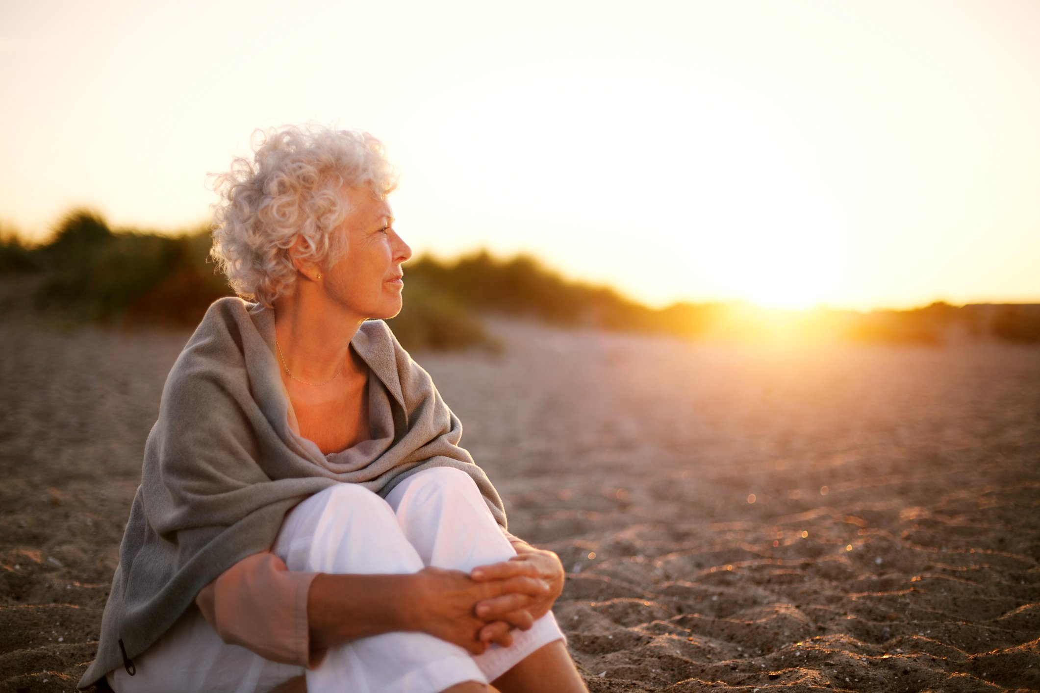 A retired woman sits on a beach and gazes thoughtfully at the sunset.