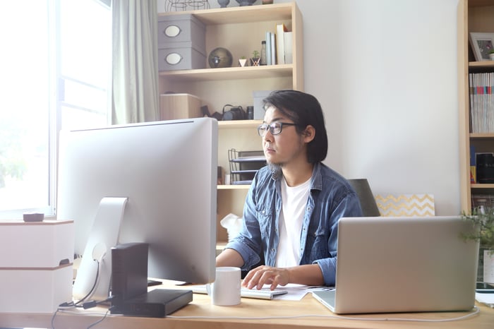 A person at a desk using a desktop computer with a laptop also open on the desk.