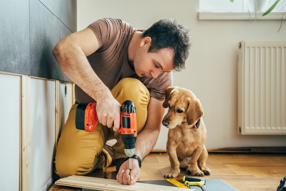 A man uses a cordless screwdriver while his puppy looks on