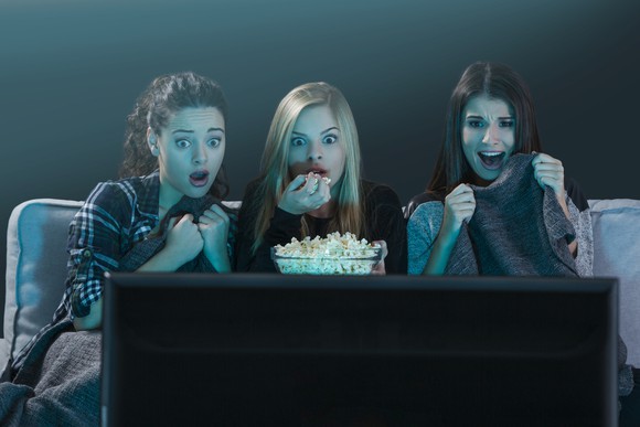 Three young women react to a scary movie on their TV.