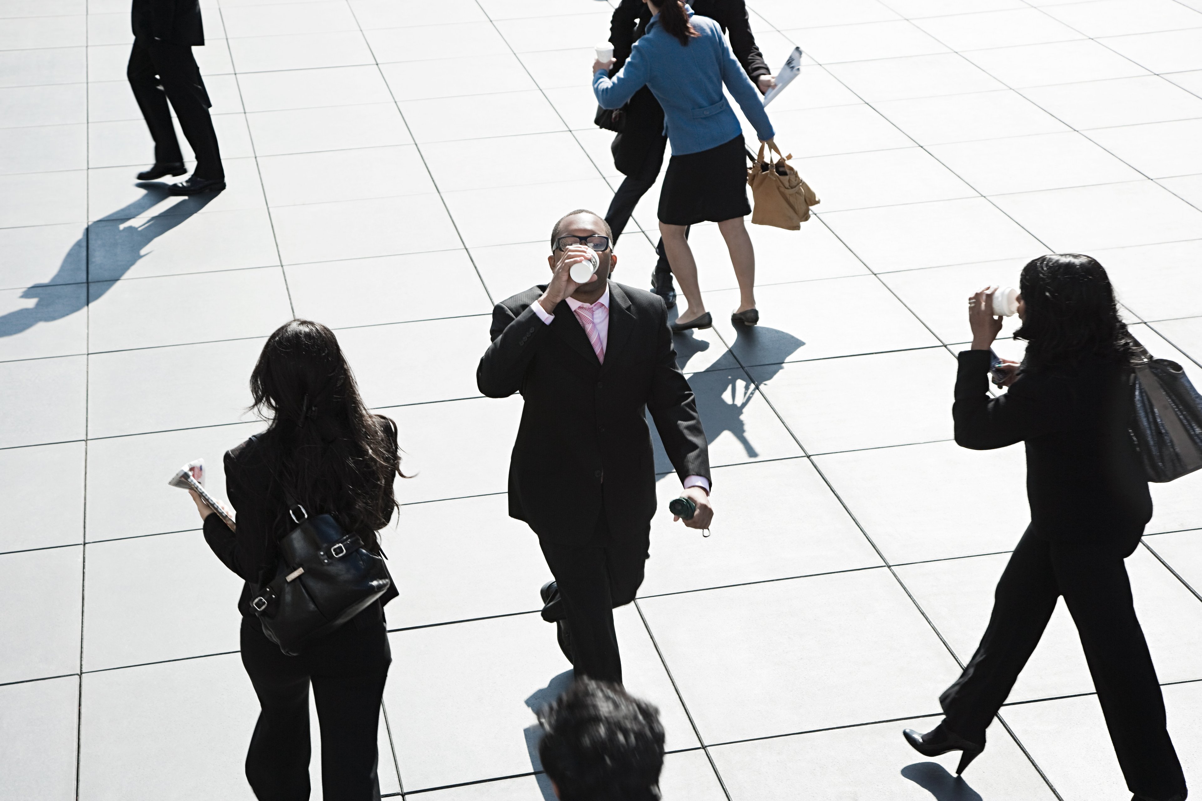 A businessman drinks a cup of coffee while walking through a crowd.