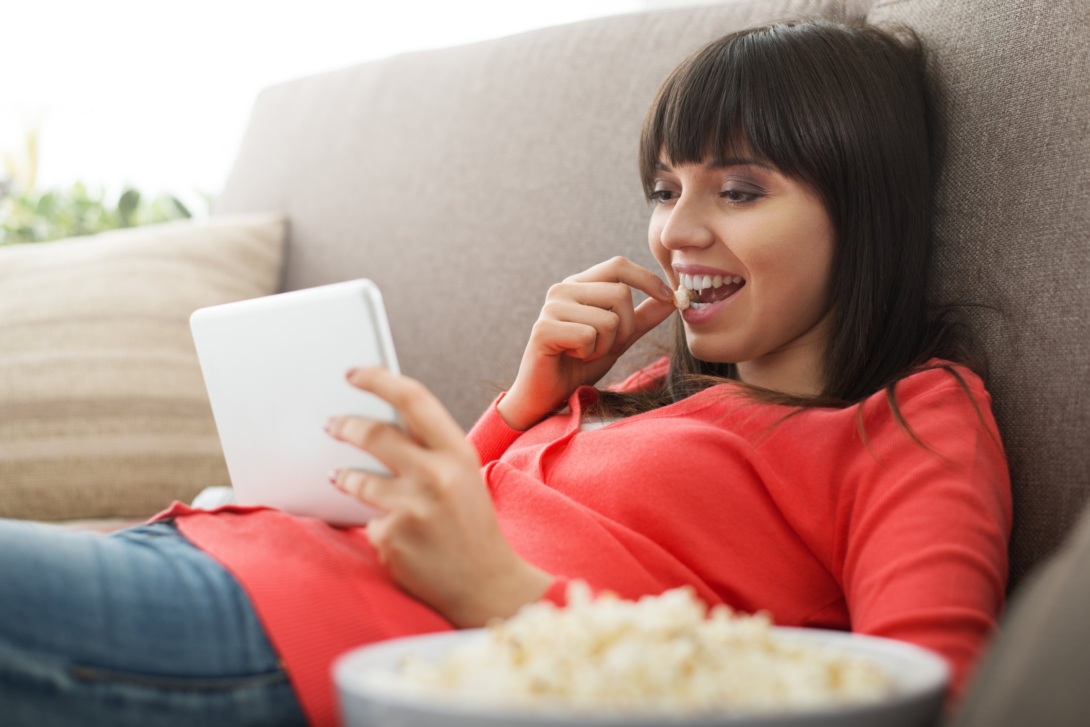 Woman sitting on couching, eating popcorn and watching a tablet