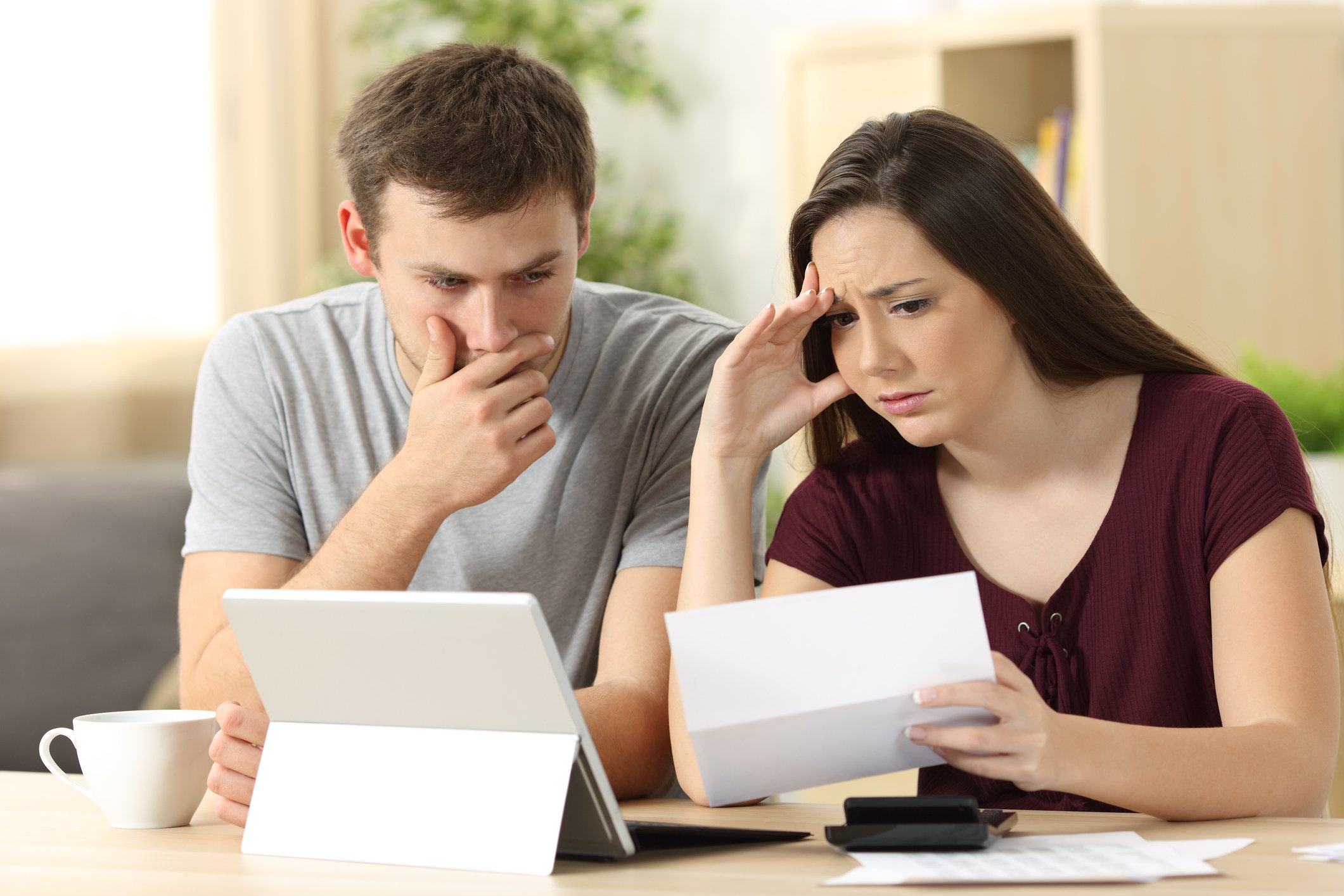 A young worried man and woman looking at a bill, with a tablet, calculator, and coffee cup on the table in front of them