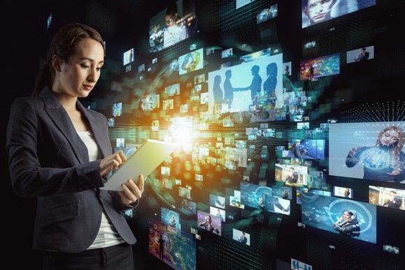 A woman with a tablet stands in front of a bank of virtual TV screens.