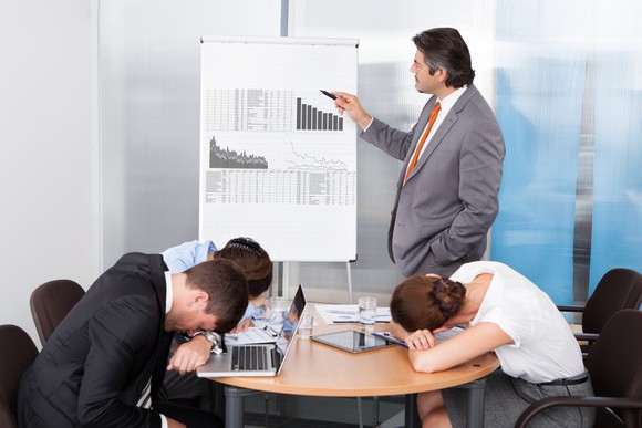 Man in suit pointing to charts while people sleep at the conference table.