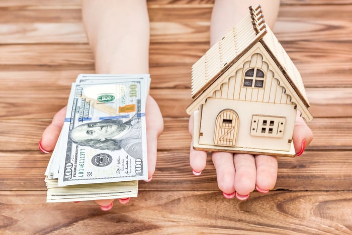 Two hands extended, with cash in one and a small model of a house in the other