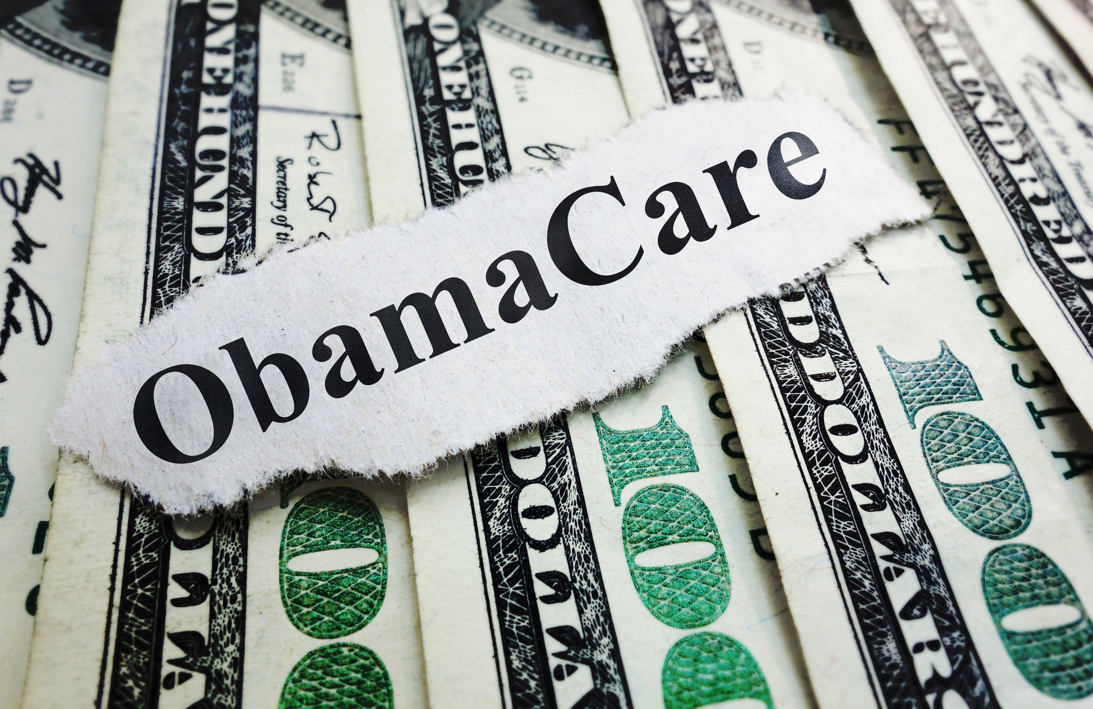 A strip of paper bearing the words "ObamaCare" rests on several hundred-dollar bills.