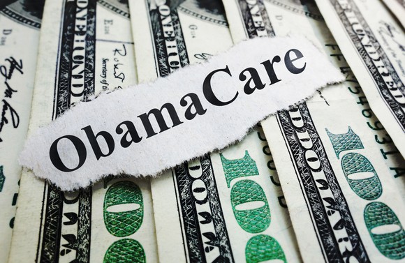 A strip of paper bearing the words "ObamaCare" rests on several hundred-dollar bills.