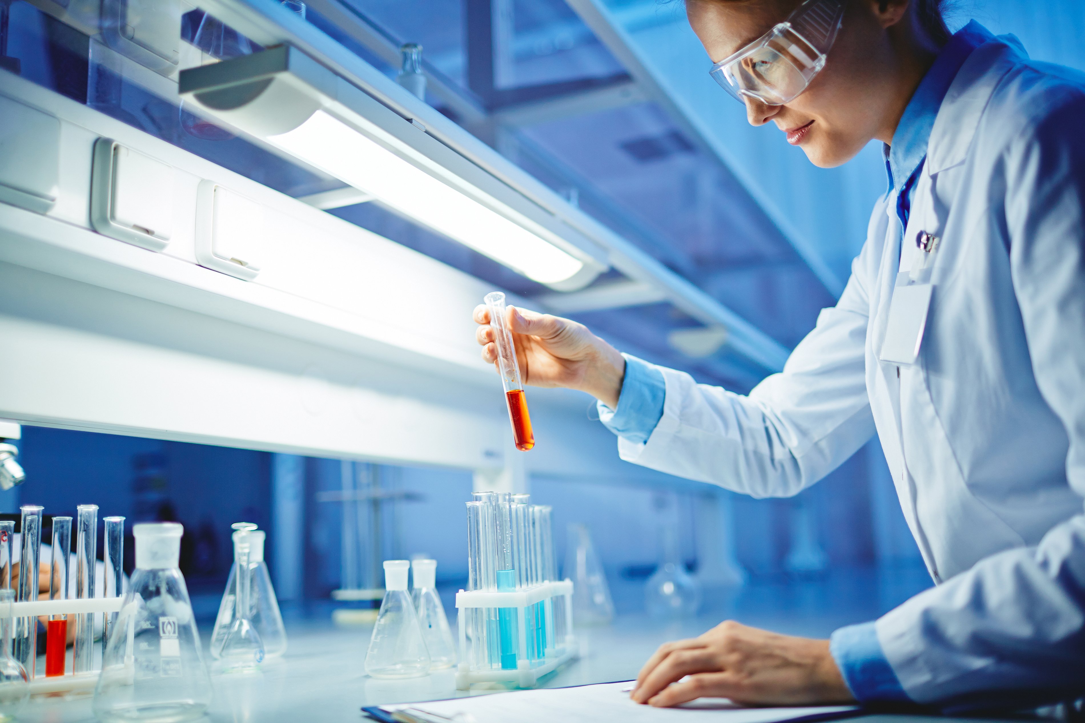 Woman in white coat and safety glasses, examining vials on a table in a laboratory