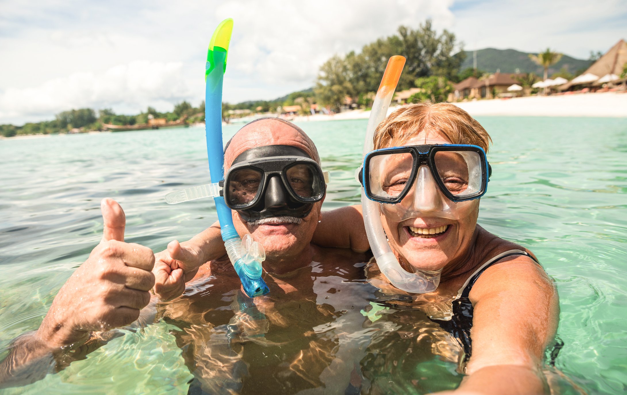 An elderly couple smiles while snorkeling in the ocean.