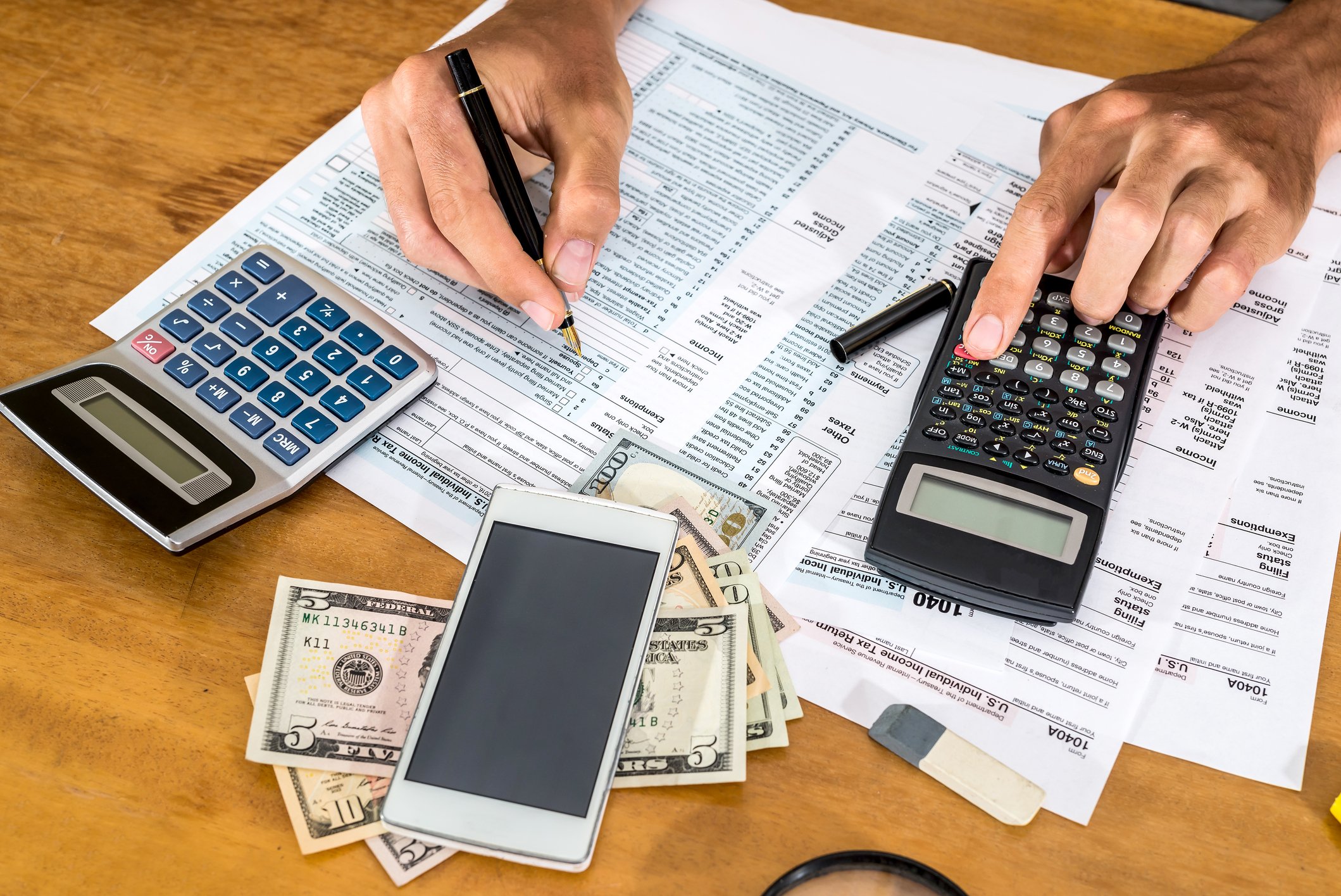 On a desk: Calculators, cash, a smartphone, and two hands writing on tax forms