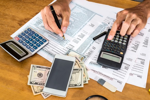 On a desk: Calculators, cash, a smartphone, and two hands writing on tax forms