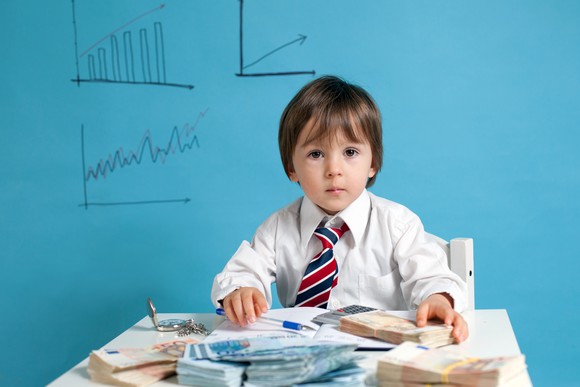 A young boy in a shirt and tie sits at a table covered with papers and cash, with charts drawn on the wall behind him.