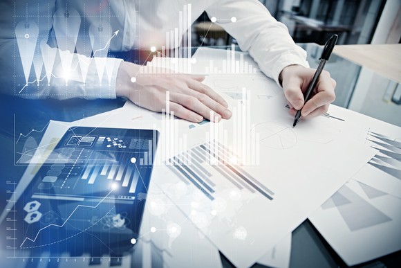 A person's arms, pen in hand, leaning over a stack of papers with bar graphs on them.