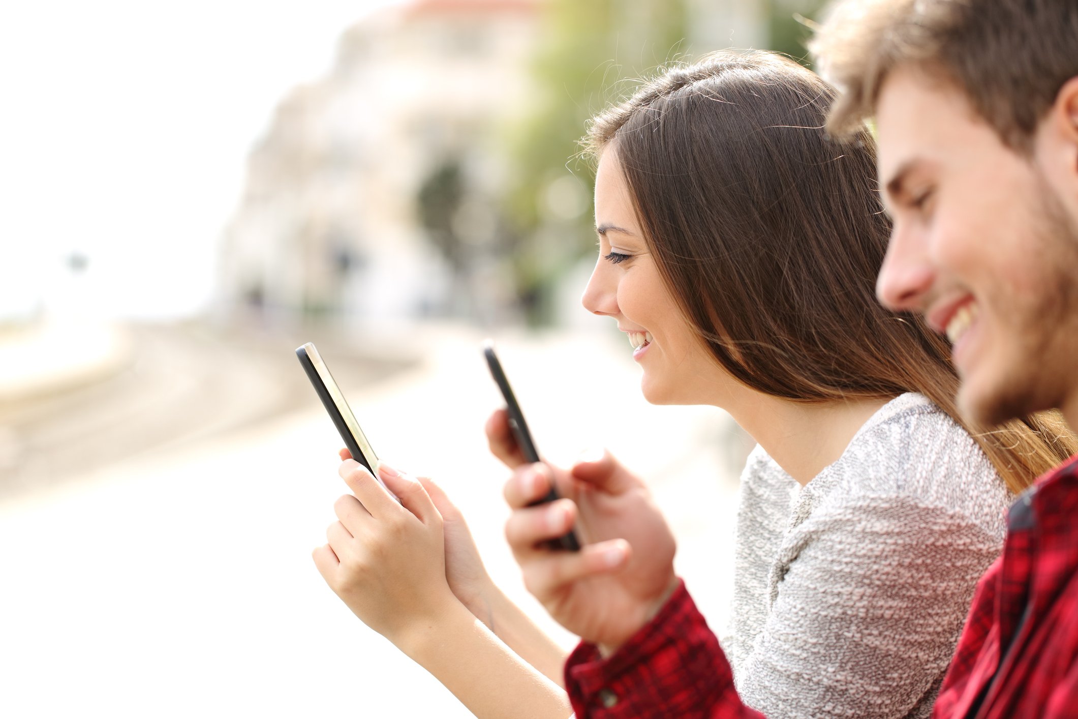 A young man and woman sit next to each other smiling at their mobile phones.