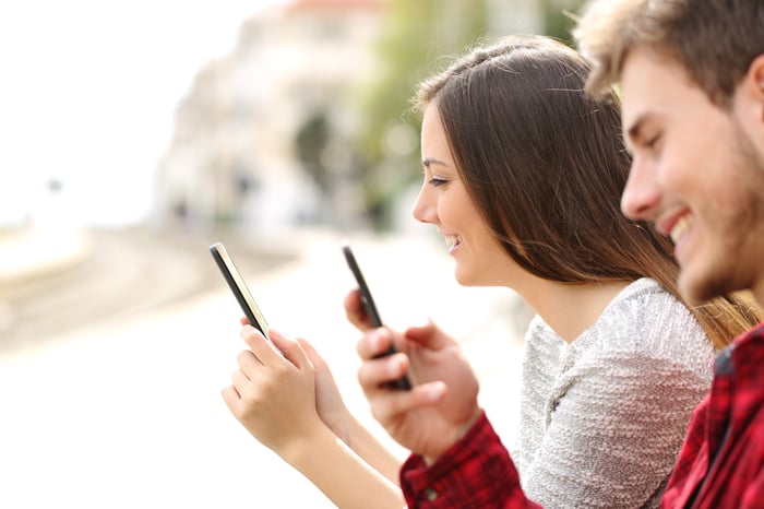 A young man and woman sit next to each other smiling at their mobile phones.