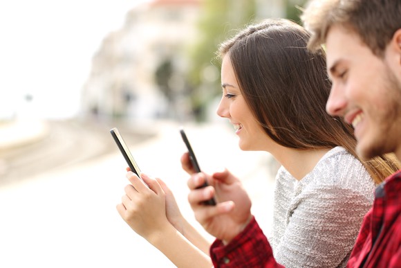 A young man and woman sit next to each other smiling at their mobile phones.