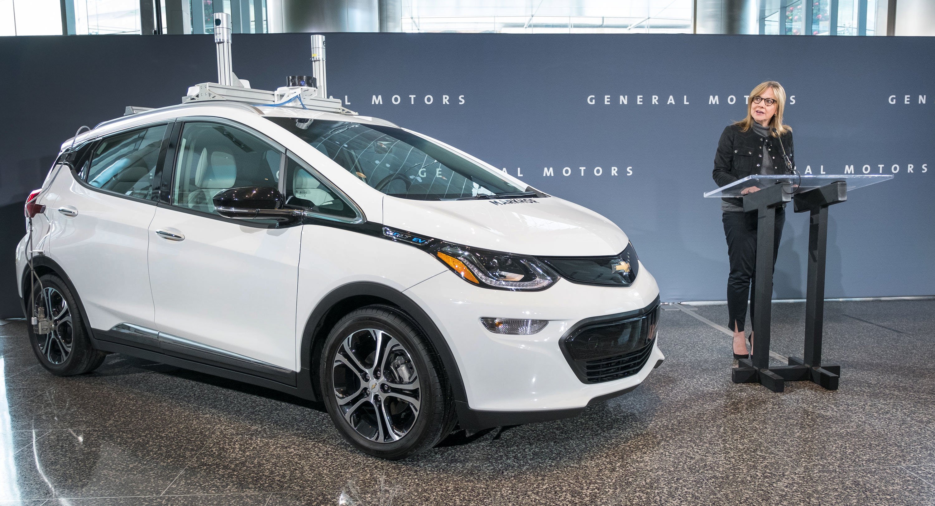 Mary Barra stands next to a white Chevy Bolt with self-driving hardware at GM headquarters in Detroit. 