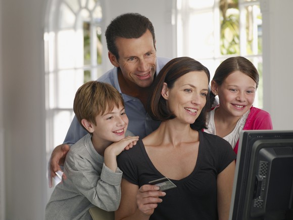 A woman sits at a computer monitor with credit card in hand, while her husband, son, and daughter huddle around her, looking at the screen