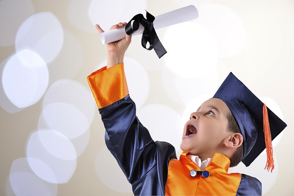 A child in a cap and gown holding up and admiring a diploma. 