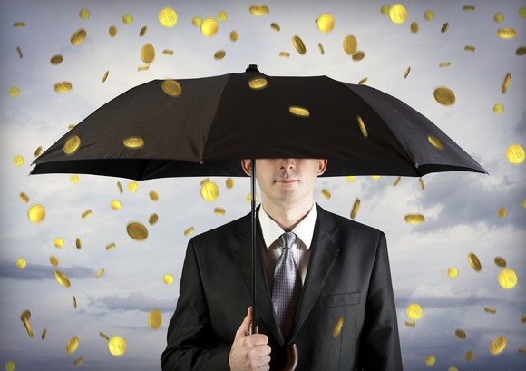 Man in suit holding an umbrella as gold coins fall out of the sky.