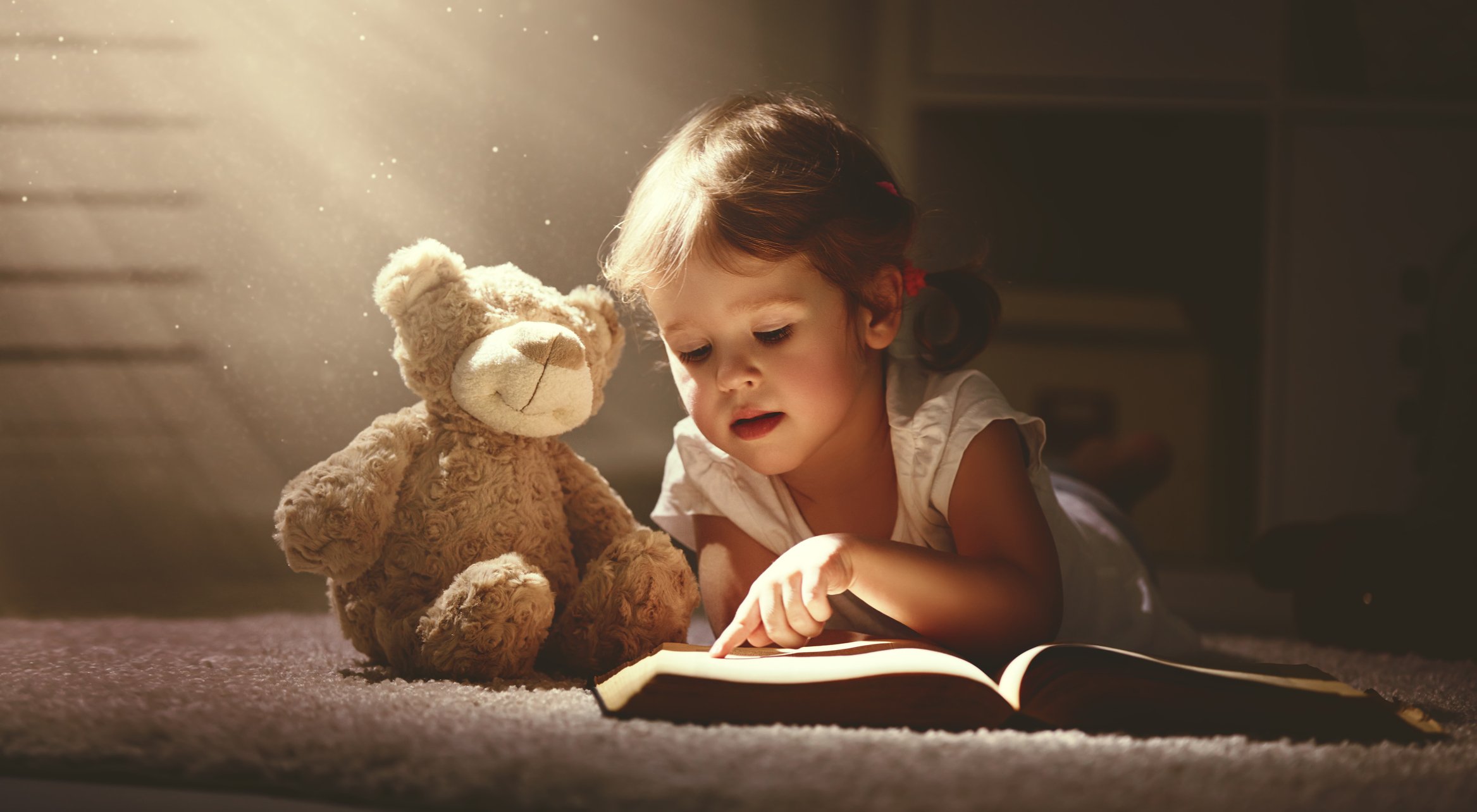 A young girl reading a book next to her teddy bear.