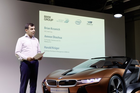 Amnon Shashua is shown standing on a stage next to a brown BMW i8 sports car.