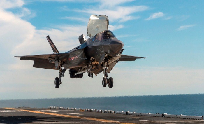 F-35B fighter jet hovering over aircraft carrier deck.