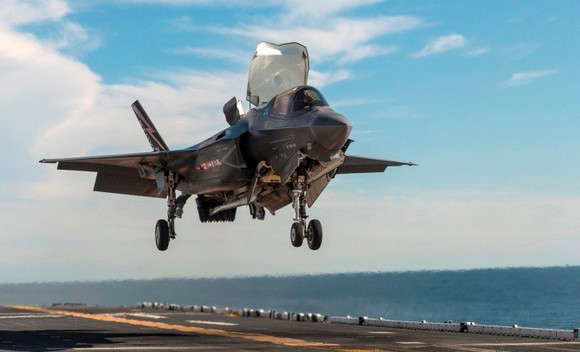 F-35B fighter jet hovering over aircraft carrier deck.