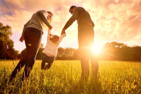 A man and a woman holding their child off the ground in a meadow at sunset.