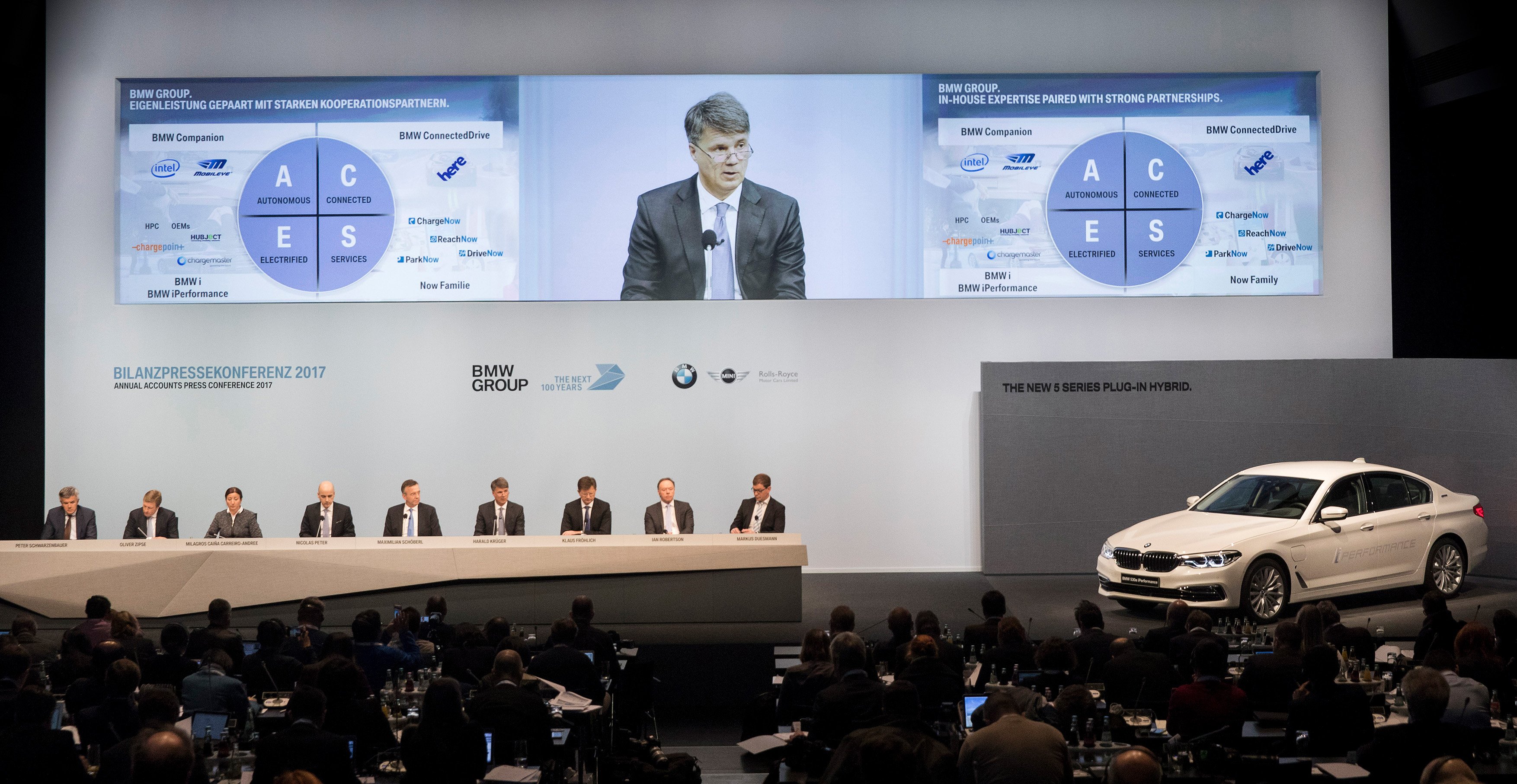 Several executives are seated on a big stage, along with a white BMW sedan. Video monitors above show a slide noting BMW's partnership with Intel and Mobileye. 