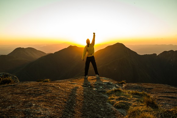 A man raising his hands in celebration, standing on a mountain top and overlooking a sunset.
