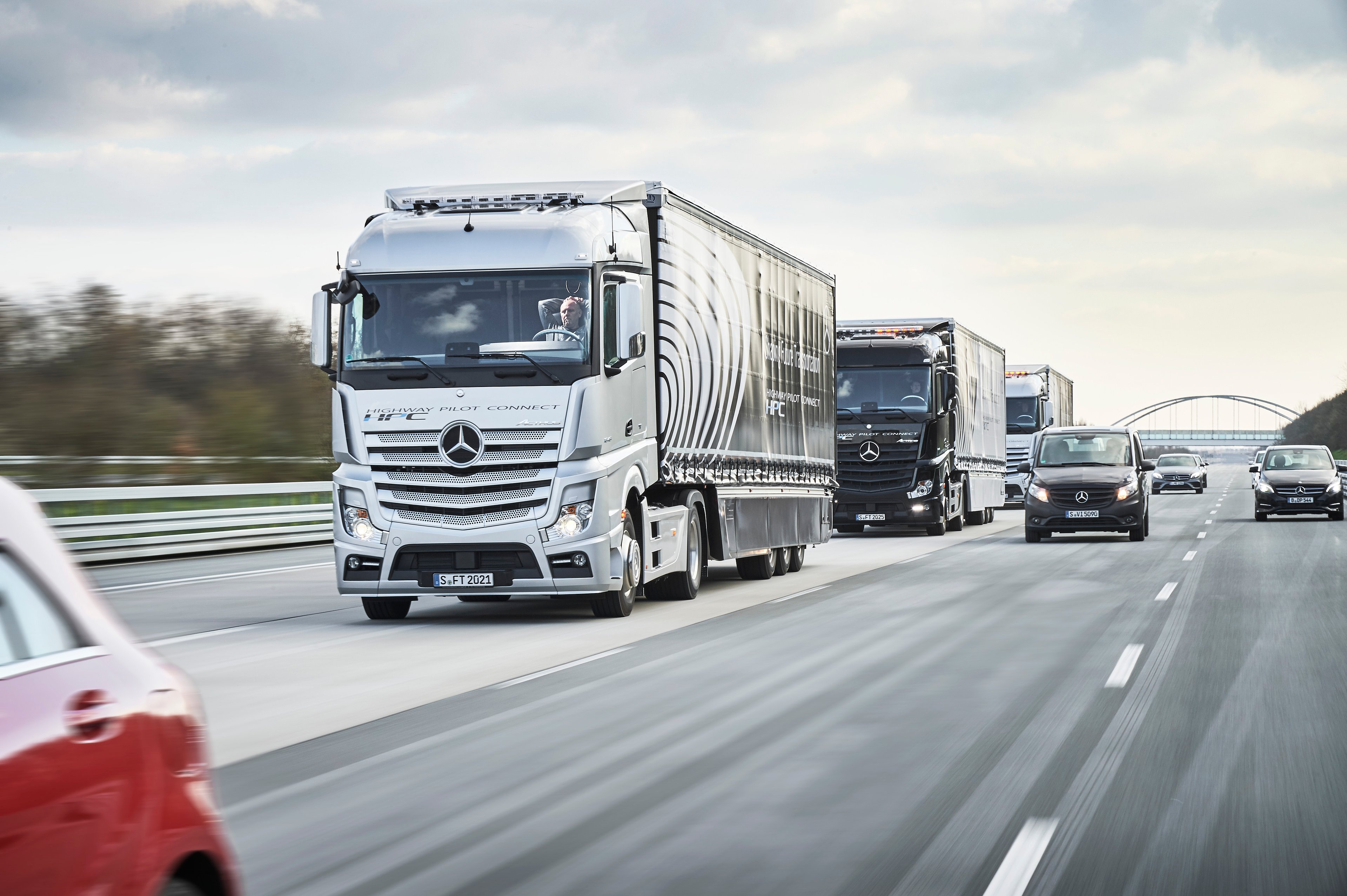 Three Mercedes-Benz tractor-trailers are shown on a highway. The driver of the lead truck has his hands behind his head, not touching the steering wheel.