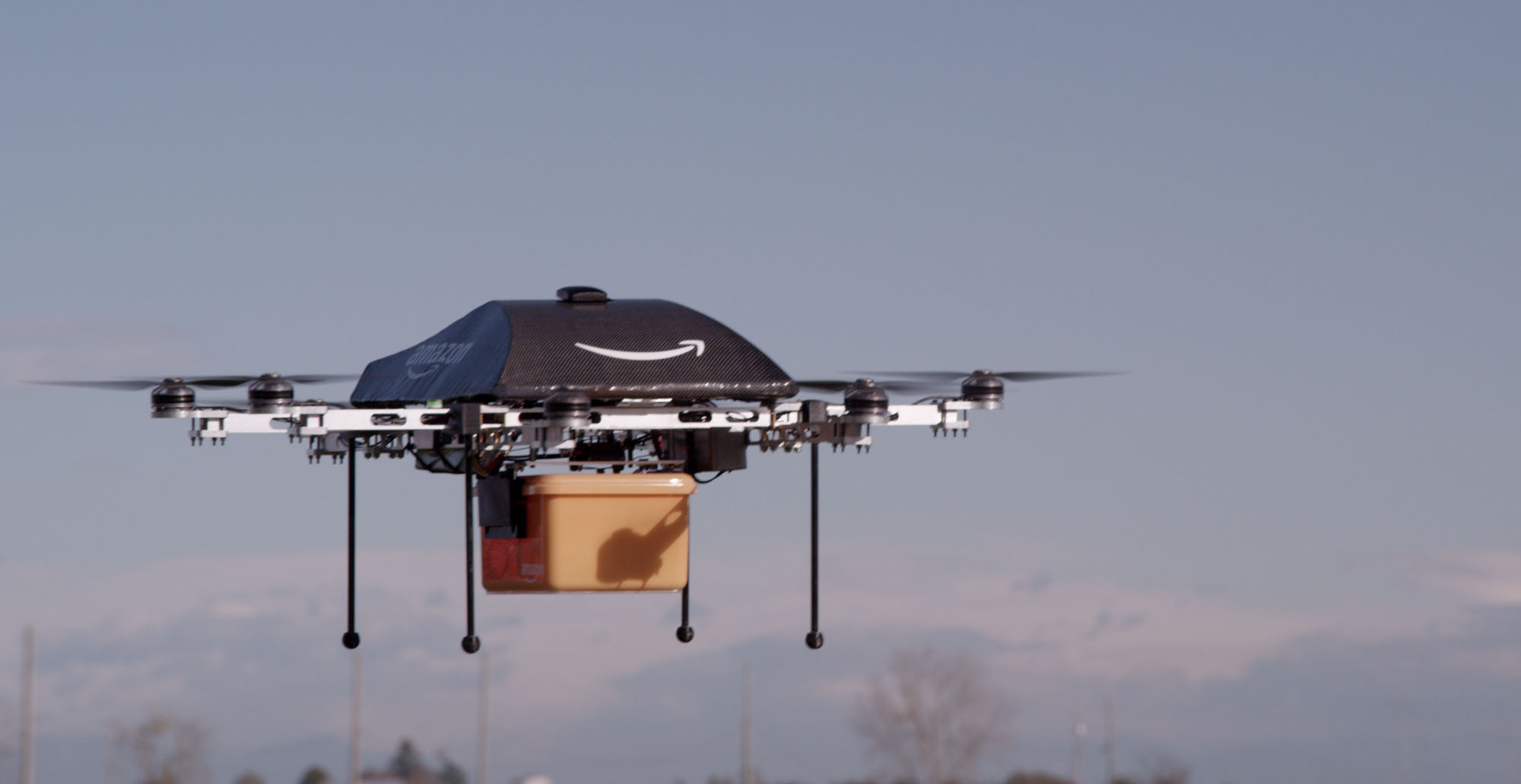 An Amazon-branded drone mid-flight holding a package in a yellow container. 