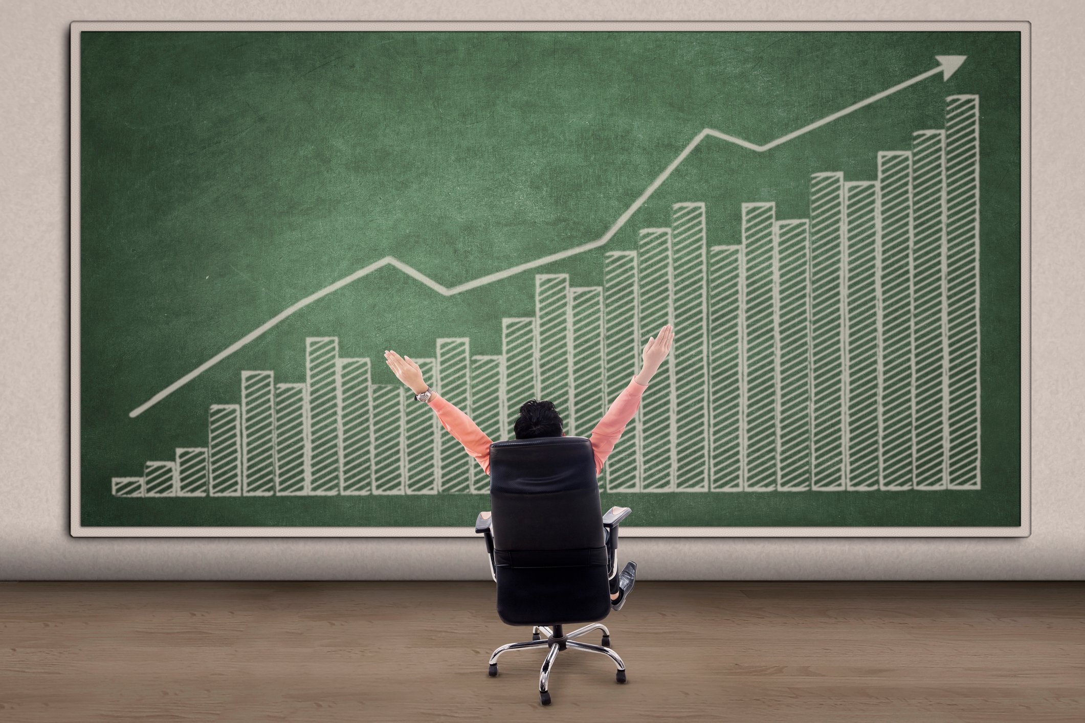A man looks at a financial chart featuring an uptrend with arms raised in celebration.