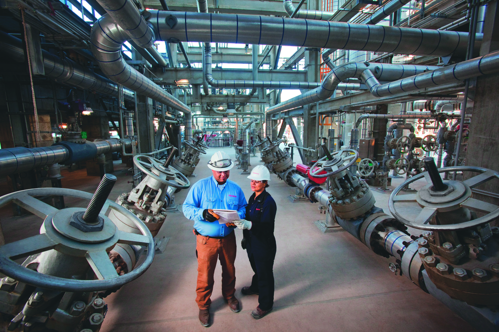 A man and woman in hard hats stand among oil pipes.