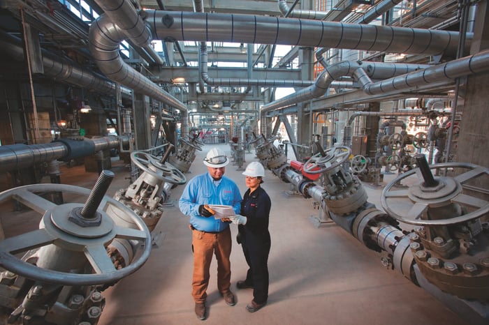 A man and woman in hard hats stand among oil pipes.