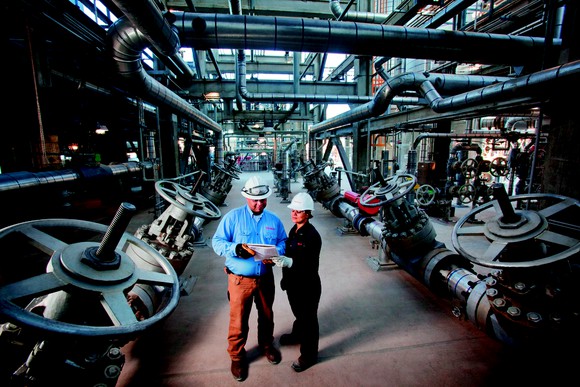 A man and woman in hard hats stand among oil pipes.