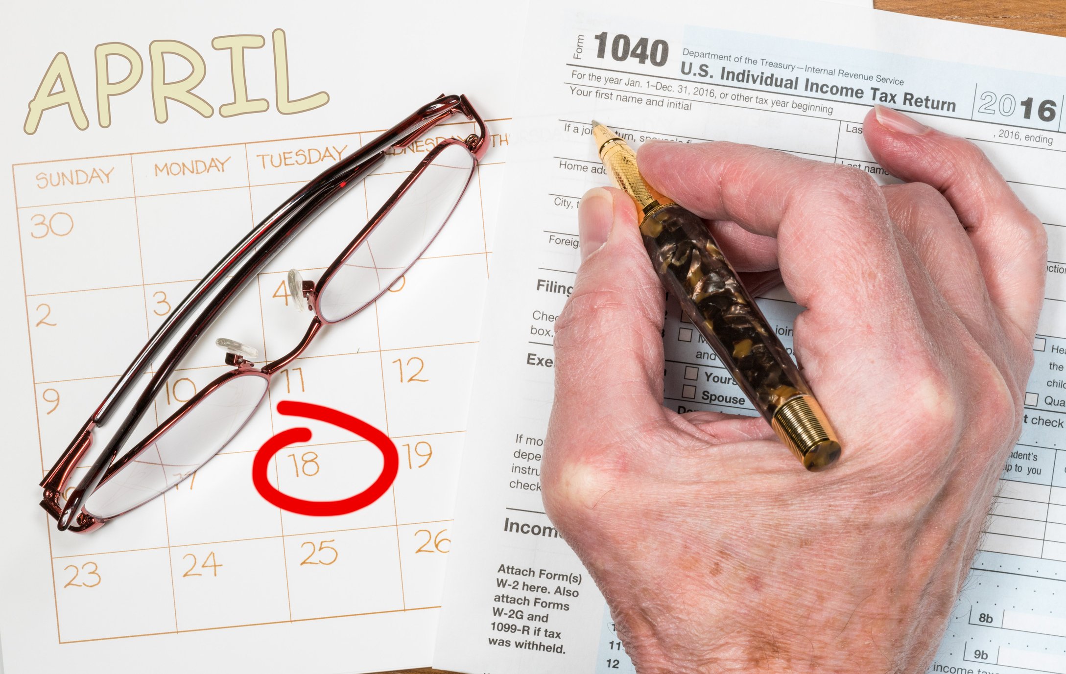 A hand holding a pen over a tax form, next to a pair of glasses and April 18 circled on a calendar.