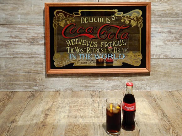 Two Coke bottles sitting in front of a old-time sign describing Coke's benefits.