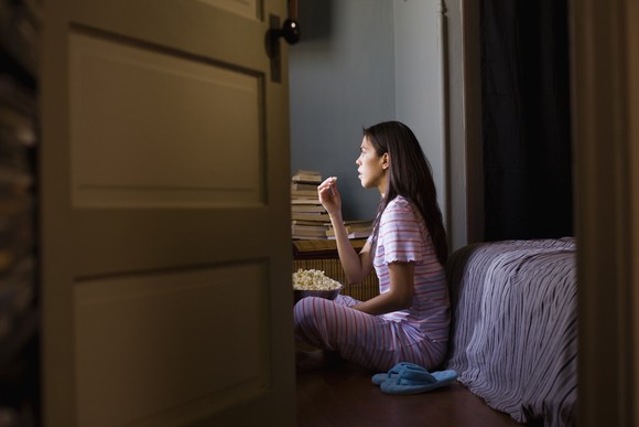 Girl watching TV while eating popcorn, seen through a half-open door. 