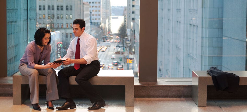 A man and a woman sit in front of a window overlooking a city while using a tablet.