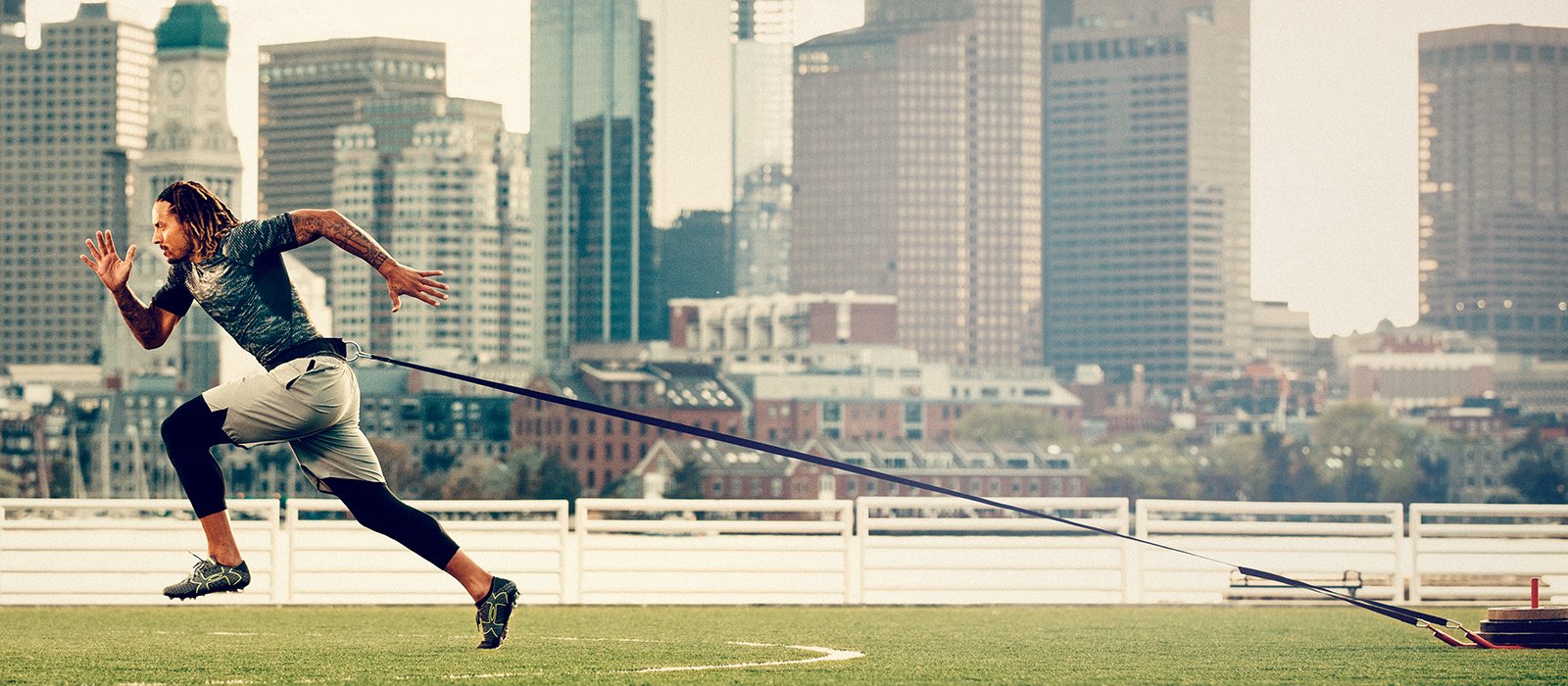 An athlete pulls a weight behind him attached to a cord while he runs across turf in Under Armour gear.