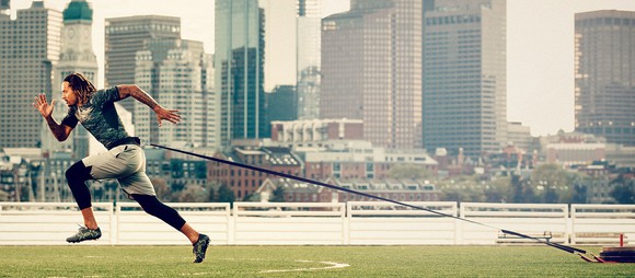 An athlete pulls a weight behind him attached to a cord while he runs across turf in Under Armour gear.