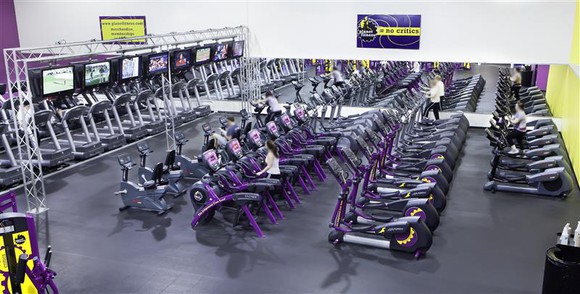 A row of exercise equipment inside a Planet Fitness. The company's "Judgement Free Zone" statement is displayed on the wall.