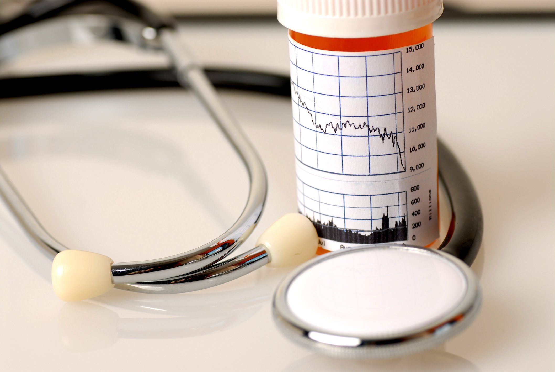 A stethoscope is wrapped around a pill bottle that has a general stock market chart on it.