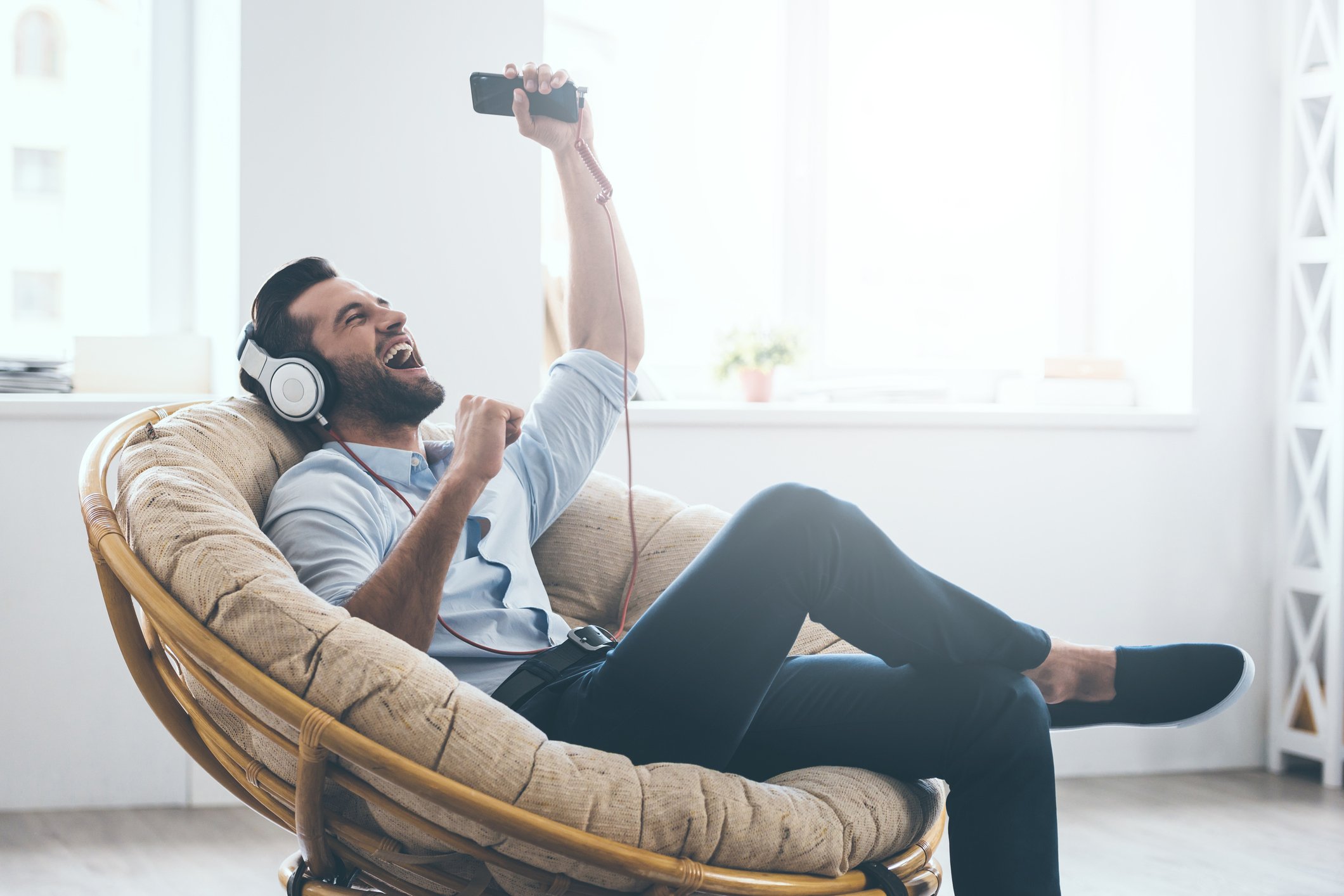 Young man in headphones gesturing and keeping eyes closed while sitting in big comfortable chair at home.