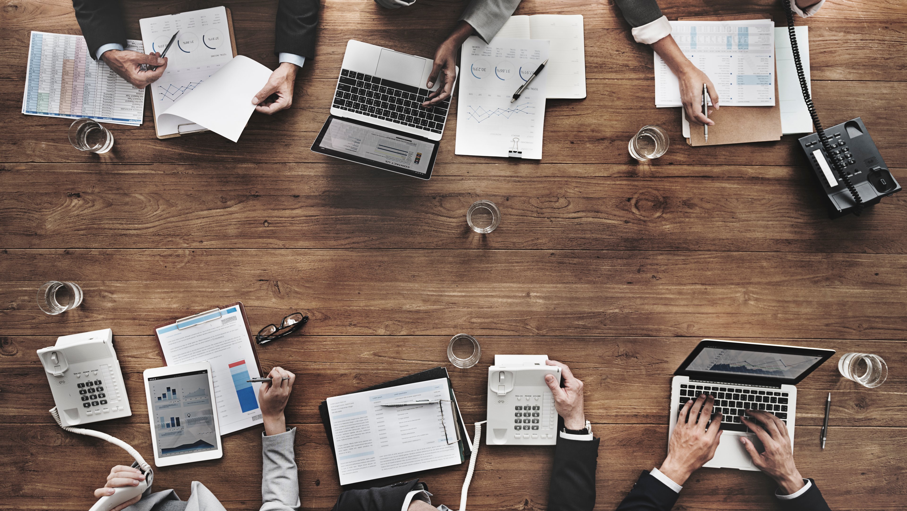 An overhead shot of a conference table, showing the hands of six businesspeople working on phones, charts, or labtops.